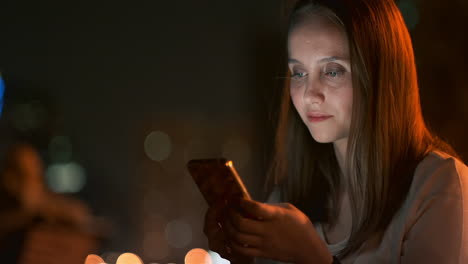 A-young-woman-in-the-night-city-looks-at-the-smartphone-screen-and-writes-a-text-message-communicates-in-social-networks-publishes-and-comments-on-photos